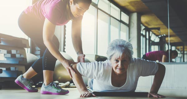 photo of an elder woman doing pushups in a gym with the help of a personal trainer